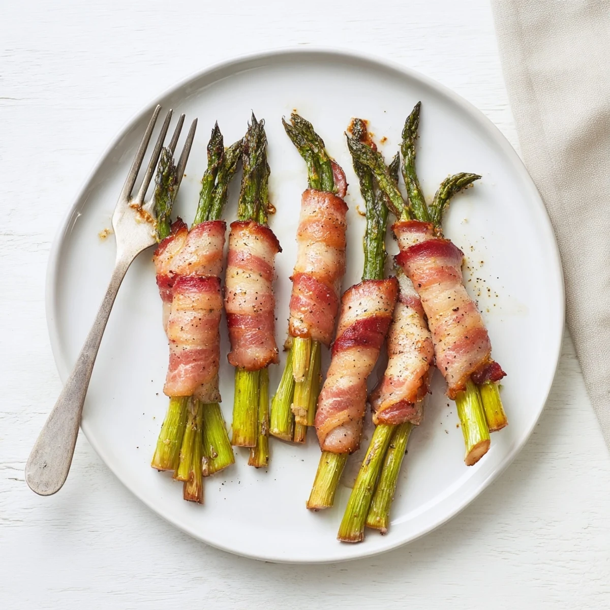 Tender green asparagus spears wrapped in crispy smoky bacon on a white baking sheet