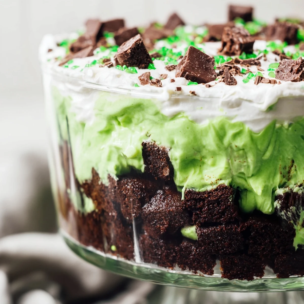 Festive St. Patricks Day brownie trifle displaying alternating layers of rich fudgy brownies, creamy green pistachio pudding, and sweet whipped cream in a clear glass dish